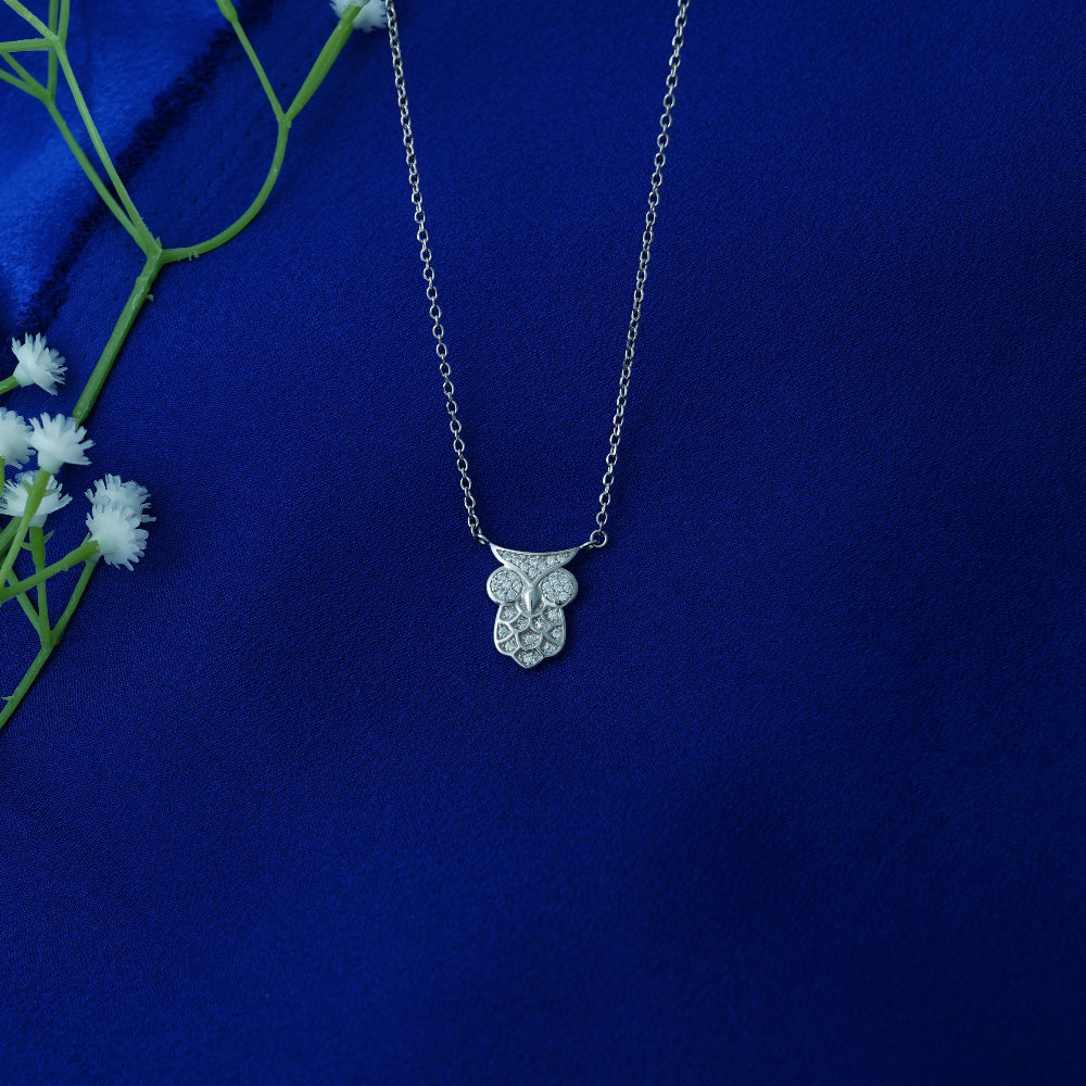 Silver owl pendant necklace on a blue fabric background with white flowers and green leaves.